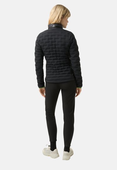 Sort isoleret jakke med quiltet design, høj krave og tætsiddende pasform. Kombineret med sorte bukser og lyse sportsko.