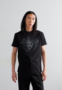 Black cotton t-shirt featuring a prominent graphic of a face and snake design in grey, with "VERSACE" text below, short sleeves, and classic fit.