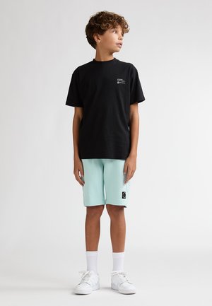 T-shirt nera in cotone con maniche corte e una piccola grafica, abbinata a pantaloncini verde menta con emblema e tasche laterali. Scarpe da ginnastica bianche e calzini.