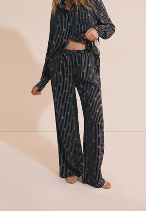 Mujer con pijama negro de rayas y estampado floral, levantando ligeramente la camisa, de pie descalza sobre un fondo beige.