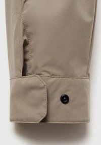 Beige stoffen jacksleeve met een gladde textuur, voorzien van een zwarte drukknoop en omgeslagen manchetten.