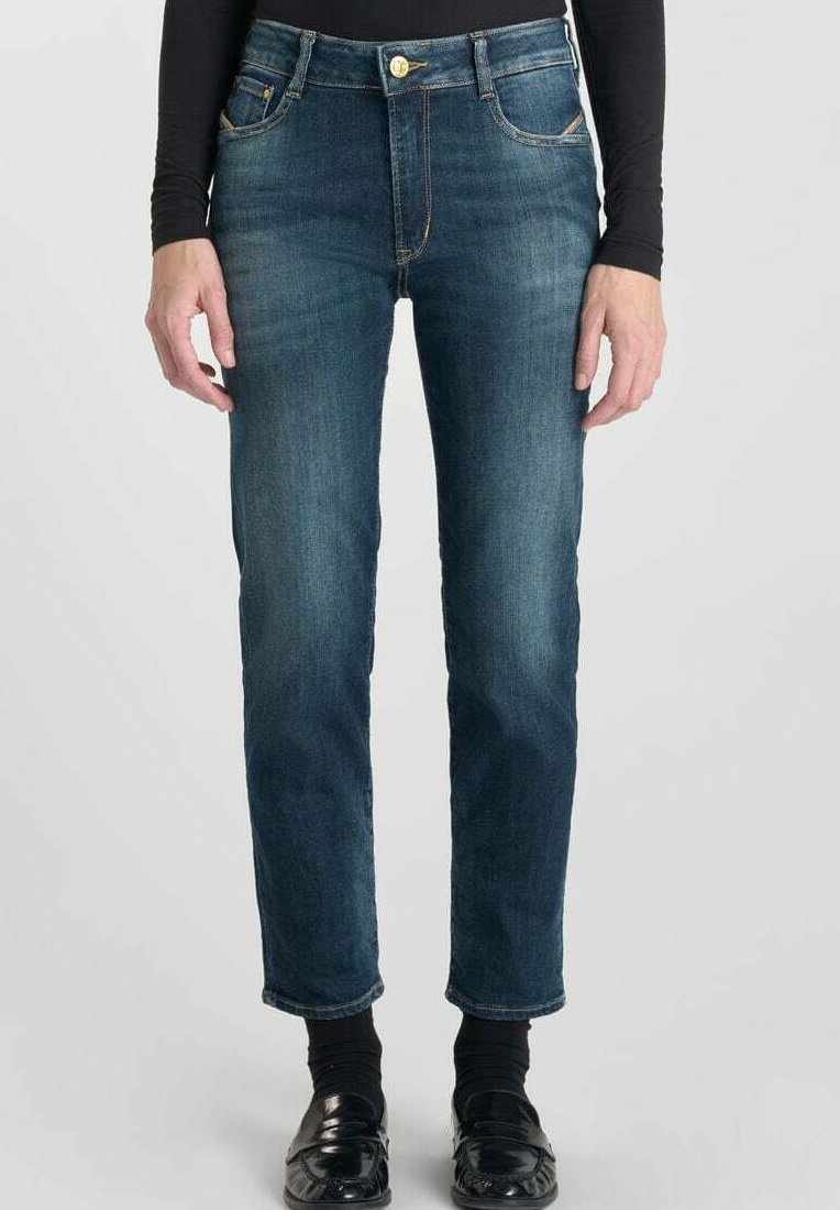 Persona vestida con vaqueros oscuros ajustados con ligeras arrugas, combinados con una blusa negra de manga larga, calcetines negros y mocasines negros, de pie.