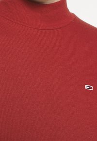Detailaufnahme eines gerippten roten Rollkragenpullovers mit einem kleinen rechteckigen Logopatch auf der Brust.