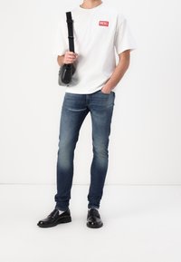 Vit bomullströja med röd "DIESEL"-logo, blå slim-fit jeans och svarta läderskor. Inkluderar en liten svart axelremsväska.