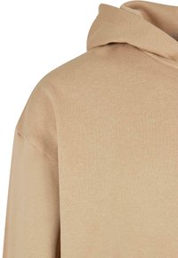 Sweat à capuche beige montrant l'épaule et le haut de la poitrine, avec un tissu doux et une coupe décontractée.