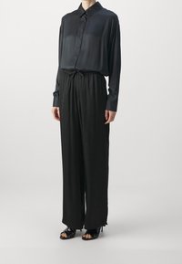 Svart satin skjorta med knappstängning och lång ärm, kombinerad med lösa svarta byxor med elastisk midja och svarta klackade sandaler med öppen tå.