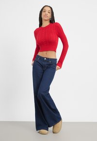 Rode gebreide cropped trui met kabelpatroon, gecombineerd met donkerblauwe flared jeans en beigekleurige enkellaarzen op een lichtgrijze ondergrond.