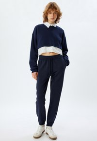 Marineblauwe cropped sweatshirt over een witte blouse met kraag, gecombineerd met marineblauwe joggingsbroek. Het sweatshirt heeft brede mouwen, de broek heeft elastische manchetten.