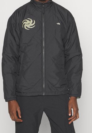 Personne portant une veste matelassée noire zippée avec un logo tourbillonnant jaune et "TNF" sur la poitrine gauche et le logo The North Face sur la poitrine droite.