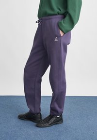 Μωβ jogger παντελόνια με ελαστική μέση και κορδόνι, που φέρουν ένα λευκό λογότυπο Jumpman στο μηρό, σε συνδυασμό με μαύρες υποδηματικές επιλογές.