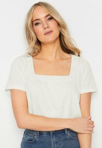 Biała koszulka z krótkim rękawem o kwadratowym dekolcie, z koronkowym wykończeniem o fakturze. Gładki materiał i luźny krój. W zestawie z dżinsami.