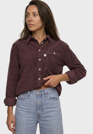 Frau trägt hochgekrempeltes bordeauxfarbenes Cordhemd und hellblaue Jeans, schaut zur Seite und hebt mit einer Hand das Hemd leicht an.