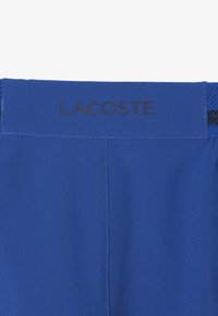 Pantaloni blu con un logo "LACOSTE" nero sulla vita, caratterizzati da una texture liscia e dettagli in rete contrastanti sui lati.