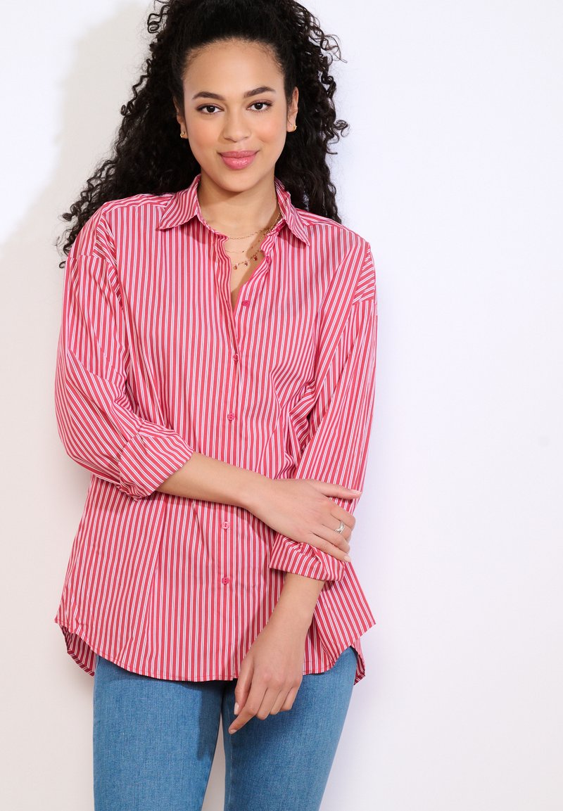 Młoda kobieta z kręconymi włosami, ubrana w czerwoną i białą koszulę w paski na guziki oraz niebieskie dżinsy, stoi na białym tle.