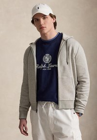 Jeune homme portant une casquette blanche, un sweat Ralph Lauren bleu marine, un sweat à capuche zippé gris clair et un pantalon blanc à cordon sur fond uni.