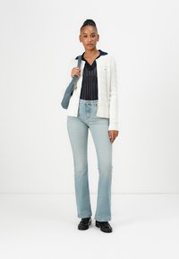 Hvit kabelstrikket cardigan, marineblå skjorte med krage, lyseblå vide jeans og svarte sko. Modell holder en grå skulderveske.
