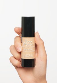 Foundationflaska med svart lock och beige kropp. Etiketten visar "Look Perfect" och "Bio-Lifting." Helt som håller den är placerad i midjehöjd.
