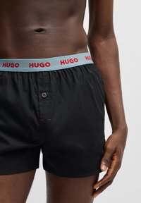 Sorte bokser-shorts med en lyseblå talje, der har et rødt "HUGO" logo. Inkluderer en knap-fly og røde kontrastsyninger.