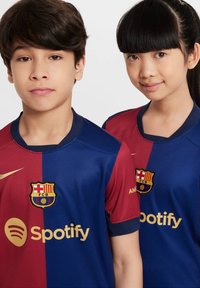 Dvoje djece nose nogometne dresove FC Barcelona, sa crveno-plavim razdvijenim dizajnom i zlatnim akcentima. Logo i sponzor su vidljivi.