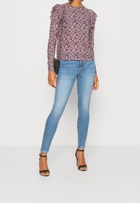 Langarmoberteil mit floralem Muster in lila und rosa, mit Rüschen an den Schultern, kombiniert mit hellblauen Skinny-Jeans und schwarzen Absätzen.