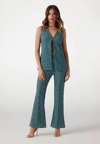 Tyrkysový vzorovaný top bez rukávov s hlbokým výstrihom do V a zodpovedajúce rozšírené nohavice. Tkanina má textúrovaný povrch; obe časti sú strihané na priliehavý vzhľad.