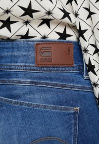 Blå denimjeans med en beige läderlapp märkt "RAW." Tyget har en slät yta med synlig sömnad och bakfickor.