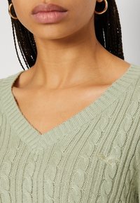 Hellgrüner, gerippter Strickpullover mit V-Ausschnitt und strukturiertem Kabelmuster. Verfügt über ein kleines Logo auf der Brust und eine weiche, atmungsaktive Textur.