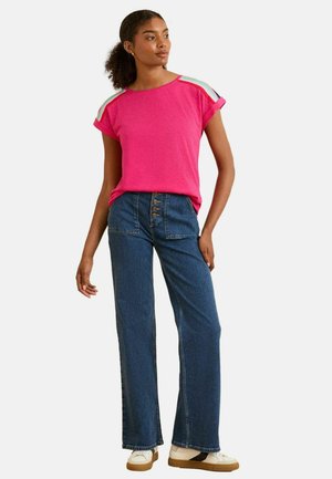 Rosa T-Shirt mit kurzen Ärmeln und aufgerollten Manschetten, kombiniert mit hochgeschnittenen blauen Jeans mit Schlag, die einen Knopfverschluss und seitliche Taschen haben.