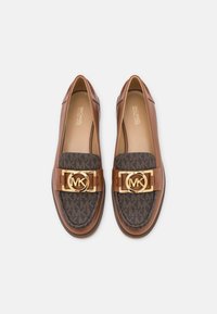 Bruine leren loafers met donker gemusterde stof, gouden ronde MK-logo gespen en beige binnenzolen met het label Michael Kors, bovenaanzicht op wit.