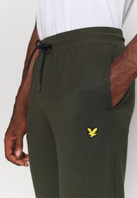 Groene katoenen joggers met elastische tailleband en verstelbare trekkoord, voorzien van een geel adelaarlogo op de linker dij en zijzakken.
