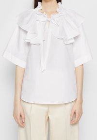 Blouse blanche avec col et manches à volants, détail noué au niveau de l'encolure ; tissu lisse et léger ; coupe ample.