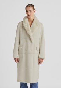 Beige Wollmischungsmantel mit doppelter Knopfreihe, breitem Kragen und zwei Vordertaschen, mit einer weichen, strukturierten Oberfläche.