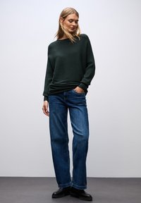 Grüner gerippter Pullover mit überschnittenen Schultern, kombiniert mit lockeren blauen Jeans. Schwarze Ankle Boots runden das Outfit ab. Neutraler Hintergrund.