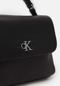 Czarna torba na ramię z teksturowanej skóry, zapaśaliowana srebrnym logo CK, klapą zamykającą i regulowanym paskiem.