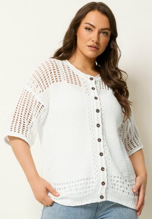 Vrouw met lang bruin haar die een witte gebreide, kortärmelig vest met knopen draagt en lichtblauwe spijkerbroek, handen rustend vlak bij de zakken.