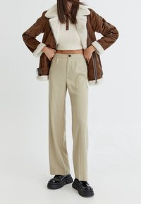 Chaqueta marrón forrada de borreguillo con cremallera plateada, top corto blanco con ribete, pantalones amplios de rayas beige y zapatos de plataforma negros.