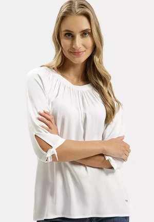 Biała bluzka z marszczonym dekoltem, rękawami o długości 3/4 z wstążkami i gładkiego materiału. Modelka pozująca z założonymi rękami.