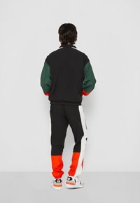 Träningsoverall i svart, grön, orange och vit. Har en zip-jacka med elastiska ärmar och matchande byxor med sidosträck och fotledsmuddar.