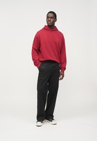 Felpa con cappuccio rossa in cotone con tasca frontale e logo minimale, abbinata a pantaloni neri e sneakers con suola bianca. Il modello è in piedi davanti a uno sfondo semplice.