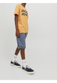 Oranje katoenen t-shirt met zwarte tekst "JACK & JONES", gecombineerd met lichtblauwe opgerolde short en marineblauwe canvas sneakers, witte sokken zijn zichtbaar.