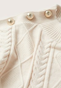 Pull color crème avec motifs torsadés texturés, col côtelé, et trois grands boutons ronds à finition métallique.