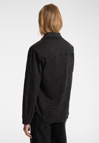 Maglietta nera con un motivo testurizzato sottile, dotata di colletto button-down e maniche lunghe. Il retro presenta un orlo curvo e dettagli minimi.