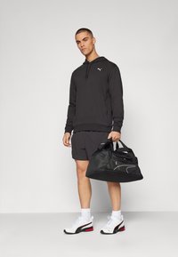 Mand iført sort hættejakke, sorte shorts og hvide sneakers, der holder en sort sportstaske mod en ensfarvet baggrund.