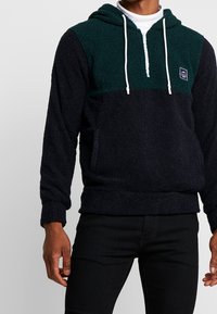Sweat-shirt en polaire dans un bleu marine foncé et un vert forêt. Il comprend une demi-fermeture éclair, une poche kangourou, des cordons de serrage blancs et un tissu texturé.