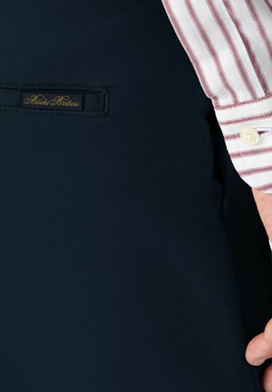 Marineblauwe broek met een soepele textuur, voorzien van een zak aan de voorkant. Een label met "Brooks Brothers" is zichtbaar, samen met een gestreepte manchette van een overhemd.