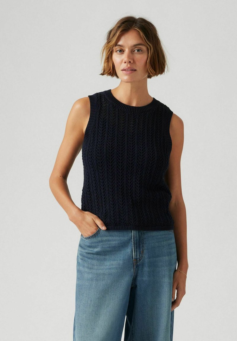 Marinblå stickad ärmlös topp med en texturerad mönstring, rund halsringning och ribbad nederkant, tillsammans med ljusblå vida jeans.