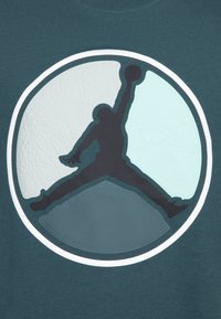 Teal t-paita, jossa on suuri, teksturoitu musta Jumpman-logo ympyrämuotoisessa suunnittelussa, jossa on vaalean vihreitä ja teksturoituja korostuksia.