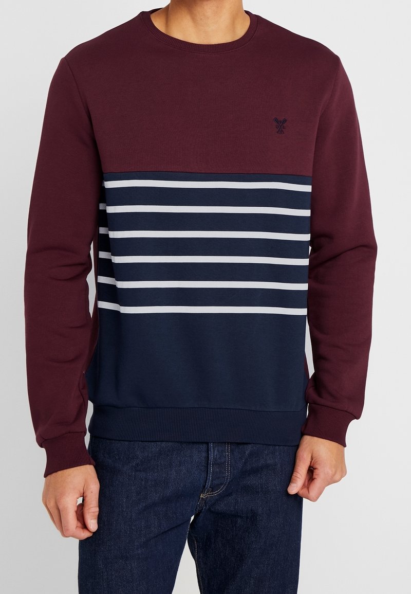 Homme portant un pull à manches longues, col rond, haut bordeaux, bas bleu marine, rayures horizontales blanches et jean bleu foncé.