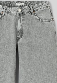 Jeans en denim gris clair avec un design classique à cinq poches, dotés d'une fermeture à bouton, d'accents cuivrés et d'une texture légèrement déchirée.