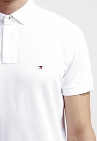 Vit polo t-shirt i strukturerad bomull med en knappslå med tre knappar och korta ärmar, försedda med en liten broderad logotyp i röd och blå.